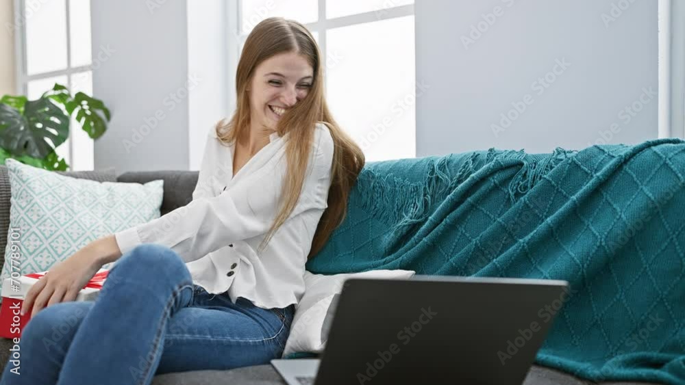 A joyful caucasian woman sitting on a couch at home, video chatting with a laptop and holding a red gift box. A joyful caucasian woman sitting on a couch at home, video chatting with a laptop and holding a red gift box.