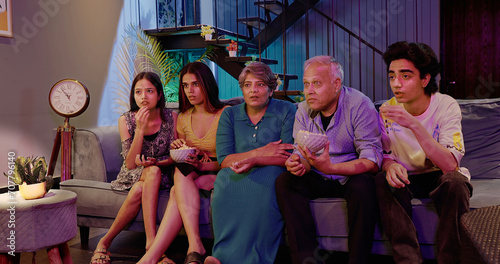 Indian Asian family friends aged mom dad man sitting sofa watch horror thriller TV scared old elder parent eat snack young adult teen girl child son hold boul people fun joy late night film movie home