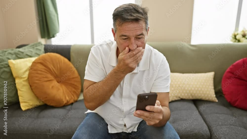 Mature man with a smartphone looks concerned in a cozy, colorful living room on a grey couch Mature man with a smartphone looks concerned in a cozy, colorful living room on a grey couch