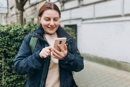 Positive cheerful woman with backpack holding smartphone gadget for communicate on the city street. 30s Woman holding on cell smartphone while relaxing outdoors, Urban lifestyle concept
