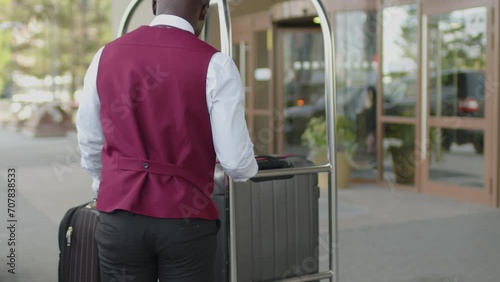 African American porter taking guests luggage from car trunk and carrying suitcase on cart into hotel photo