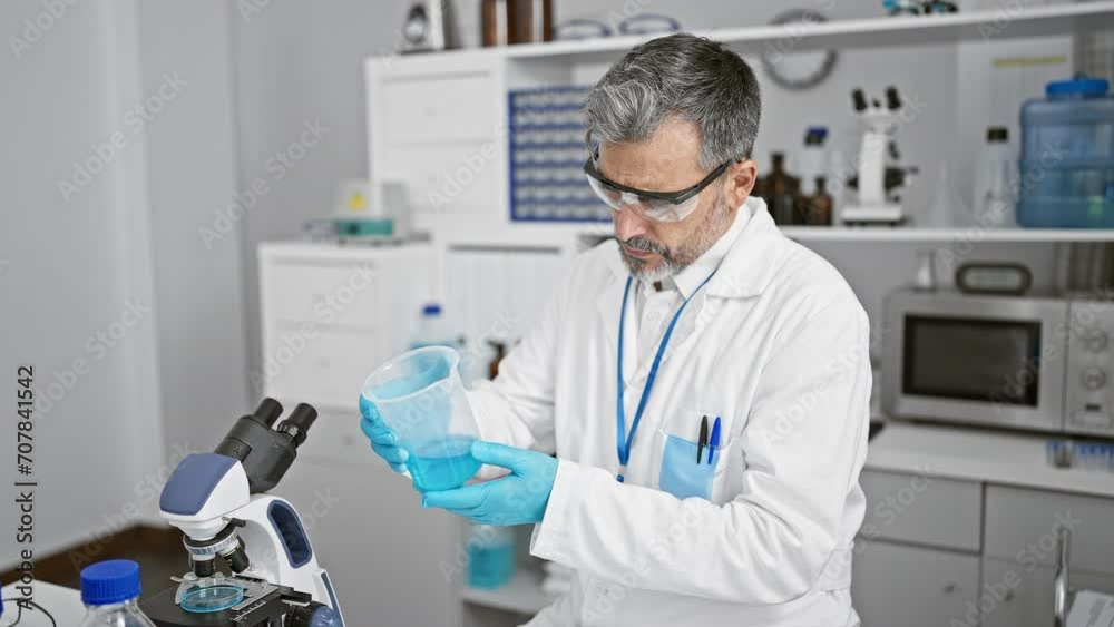 Cheerful young grey-haired hispanic man scientist confidently measuring liquid in test tube, working indoors at a busy laboratory, smiling under safety glasses and gloves Cheerful young grey-haired hispanic man scientist confidently measuring liquid in test tube, working indoors at a busy laboratory, smiling under safety glasses and gloves