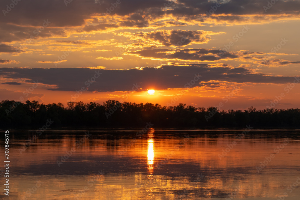 Picturesque landscape of the setting Sun surrounded by clouds in the sky, golden sunset over a wide river