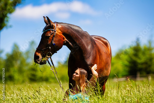A young woman rider with long brunette hair stands with her horse on a high summer meadow in the bright sunshine.