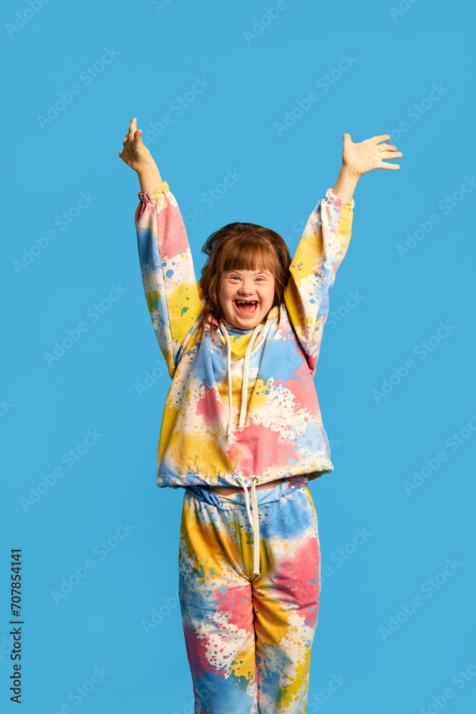 Smiling girl with dawn syndrome expressing happiness and joy against blue studio background. Tolerance, support and care. Concept of acceptance, inclusion, health, diversity, emotions - obrazy, fototapety, plakaty 