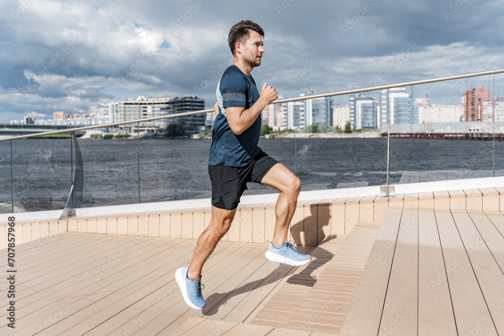 Fototapeta premium Focused man high-kneeing during an urban riverfront workout.