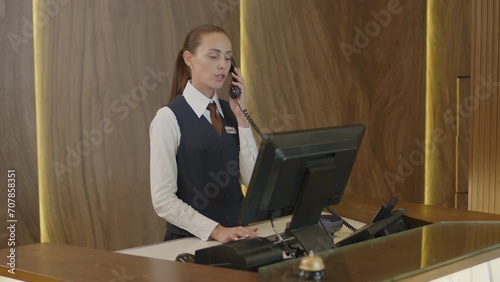 Young female hostess working on computer and answering phone call at front desk in hotel