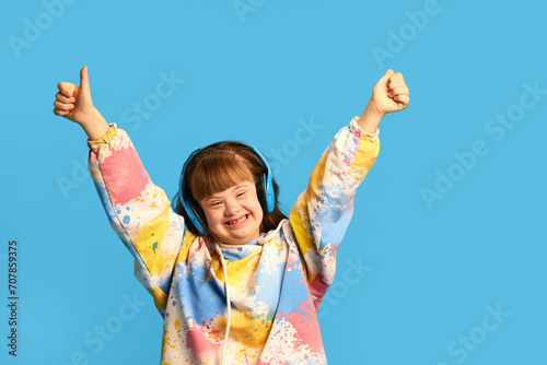 Happy smiling teen girl with down syndrome listening to music in headphones, expressing joy against blue background. Concept of acceptance, care, inclusion, health, diversity, emotions, equality photo