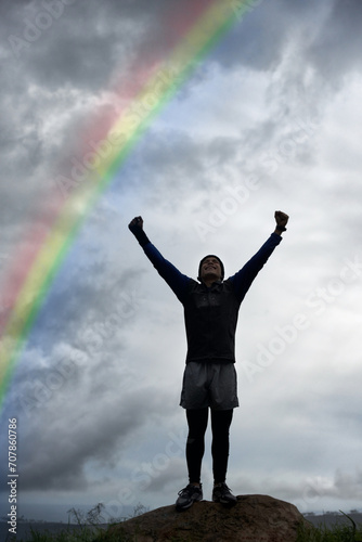 Man, rainbow and cloudy sky in celebration for freedom, achievement or fitness standing on mountain. Active male person with fist pump in victory for workout, exercise or outdoor training success