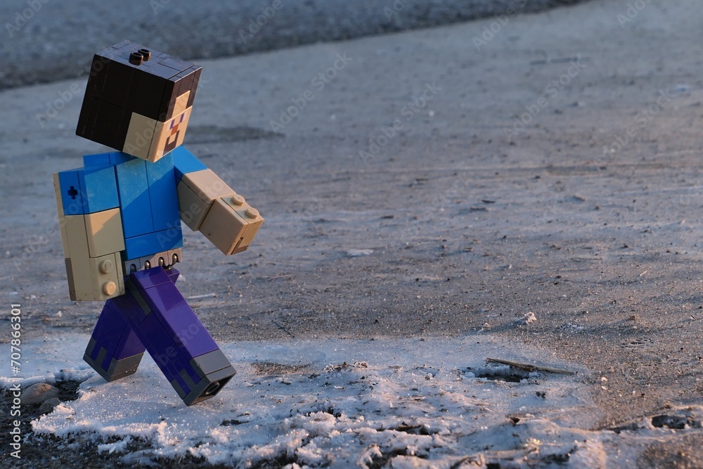 Fototapeta premium LEGO Minecraft figure of Steve walking along frozen water surface near asphalt road, winter morning sunshine.
