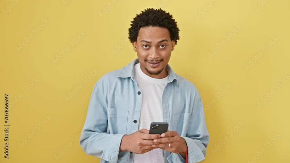 Cheery young african american man with a toothy smile, standing tall, confidently holding a smartphone - exuding a positive attitude, in front of a yellow isolated background. Cheery young african american man with a toothy smile, standing tall, confidently holding a smartphone - exuding a positive attitude, in front of a yellow isolated background.