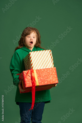 Emotional teen girl with down syndrome standing with socked face, holding many present boxes on green studio background. Concept of acceptance, care, inclusion, health, diversity, emotions, equality © master1305