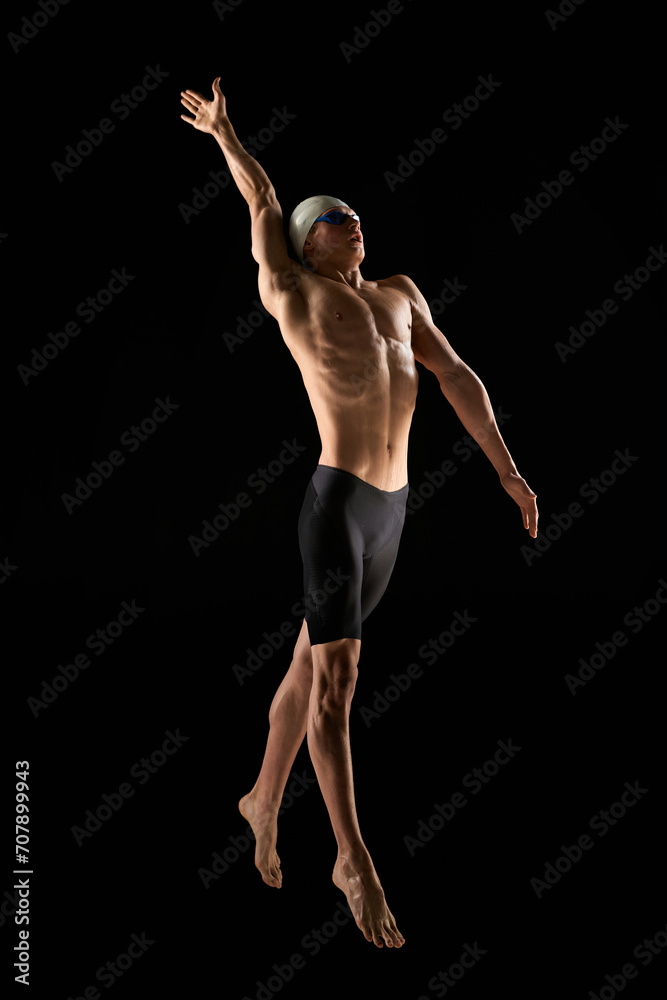 Full-length image of muscular young man, professional swimmer with muscular relief body shape training, showing swimming techniques over black background. Concept of water sports event, competition