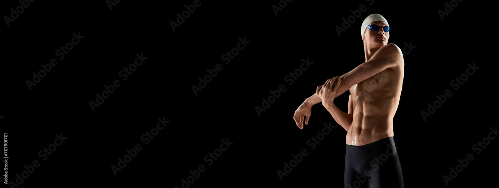 Φωτογραφία Muscular, athletic young man, swimmer in goggles and cap stretching muscular body before swim over black background