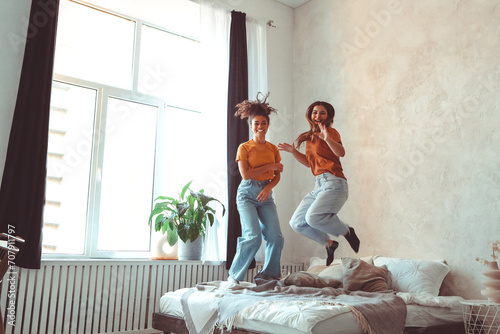 Two happy joyful girlfriends of different races jumping on bed and having fun together at home photo