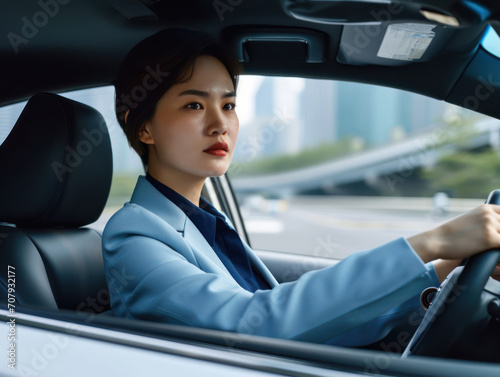 a happy stylish asian woman in light blue suit is driving white car. Portrait of happy female driver steering car with safety belt. © Svetlana