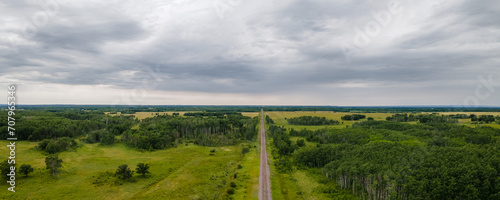 A straight railroad line runs into the distance through flat scrubby farmland with green fields and scattered trees. The sky is filled with gray clouds. 