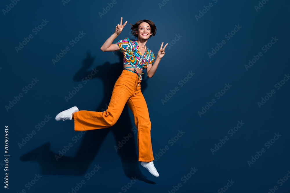 Full length photo of lovely young lady running showing double v-sign wear trendy flared pants isolated on dark blue color background Full length photo of lovely young lady running showing double v-sign wear trendy flared pants isolated on dark blue color background