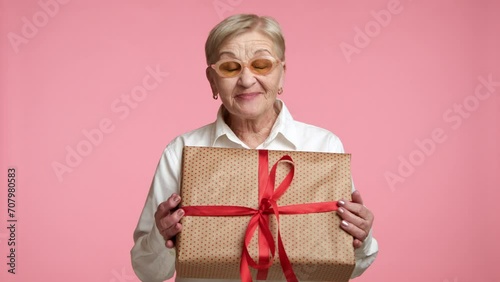 Well-groomed old lady in spectacles swoon with happiness, opening mouth with excitement, pressing present in golden paper with red ribbon, pink background. High quality 4k footage photo