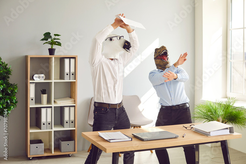 Half animal half people having fun at end of work week. Male corporate workers in horse and dinosaur masks having party. Funny happy men with weird horse and T rex dino faces dancing by desk in office photo