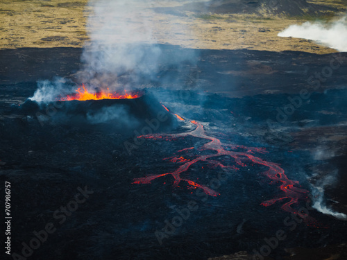Erupting volcano, red hot boiling lava pouring out of crater into the devastated surrounding area, drone shot. Earth, nature, and power concepts.