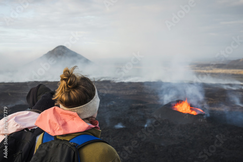 Woman tourist looking at dramatic view of dark volcanic landscape with glowing melting lava boiling in the crater, active volcano during eruption in Iceland