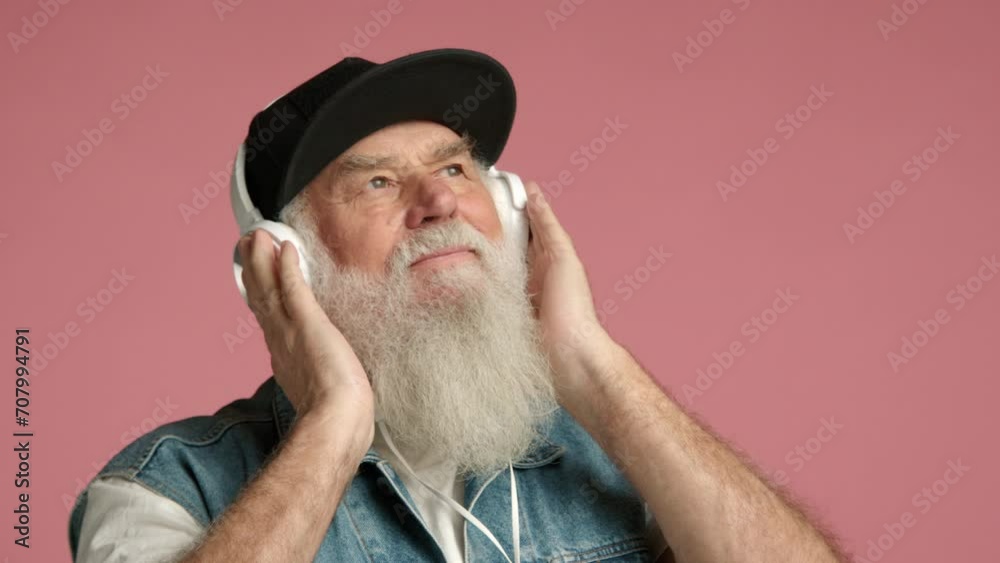 Elderly man with a long white beard wearing headphones and a black cap, enjoying music with a peaceful expression, against a pink background. Camera 8K RAW. Elderly man with a long white beard wearing headphones and a black cap, enjoying music with a peaceful expression, against a pink background. Camera 8K RAW.