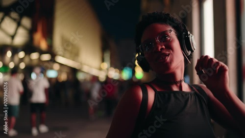 Energetic woman dancer in city, headphones delivering soundtrack to blissful carefree night. Happy female trendy technology user moves her body in rhythm of favorite son of playlist. Slow motion photo