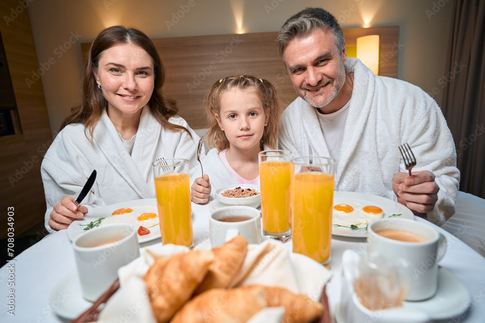 Happy caucasian family having breakfast and looking at camera in hotel room