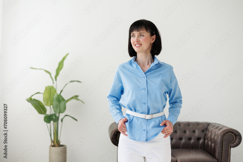 Women equality day. Smiling successful businesswoman in elegant blue shirt and white pants looks at camera at modern comfortable light office with green plant and sofa. Work life balance concept. Women equality day. Smiling successful businesswoman in elegant blue shirt and white pants looks at camera at modern comfortable light office with green plant and sofa. Work life balance concept.