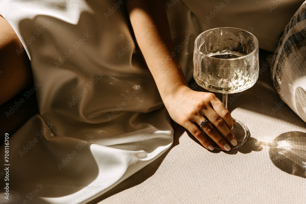 Female hand with diamond engagement ring holding a glass of champagne in sunlight. - obrazy, fototapety, plakaty 