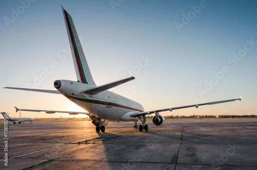 Rear three quarter view of passenger airplane on airport apron and few other planes in the background. Low evening sun creates a long shadow of aircraft silhouette on concrete surface. 