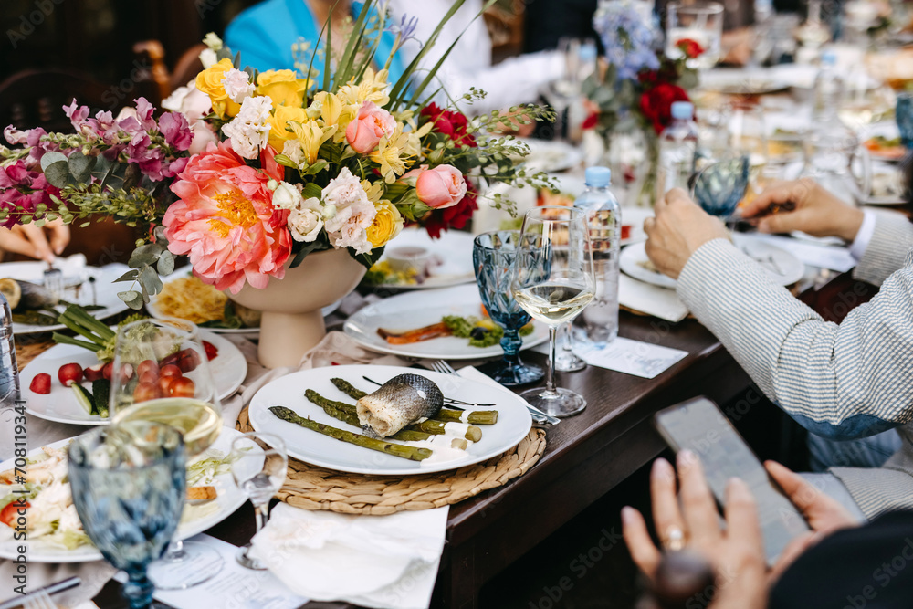 Festive table setting with colorful floral centerpiece, guests dining, fish dish, asparagus, wine, water, and vibrant atmosphere.