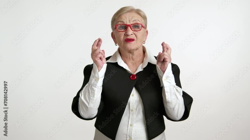 Optimistic elderly lady with red glasses crossing her fingers, making a wish or hoping for positive news against a white background. Optimistic elderly lady with red glasses crossing her fingers, making a wish or hoping for positive news against a white background.