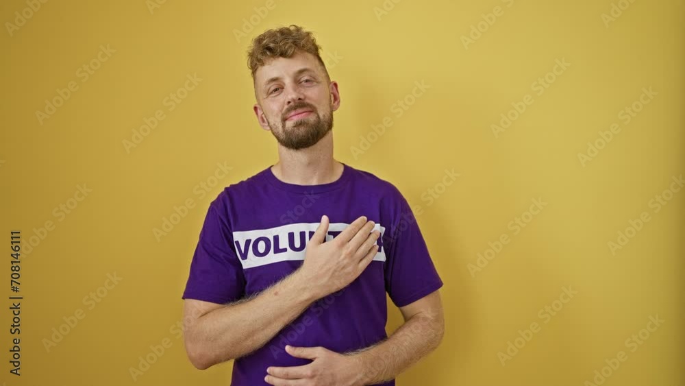 A handsome young man with blue eyes and beard in a purple volunteer shirt posing against a yellow wall A handsome young man with blue eyes and beard in a purple volunteer shirt posing against a yellow wall