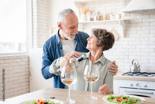 Happy old elderly senior couple spouses grandparents celebrating special event anniversary Valentine`s day at home, drinking wine, having romantic dinner date in the kitchen