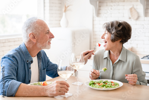 Happy old elderly senior caucasian family couple grandparents spouses treating each other while eating, celebrating anniversary on romantic date dinner at home kitchen