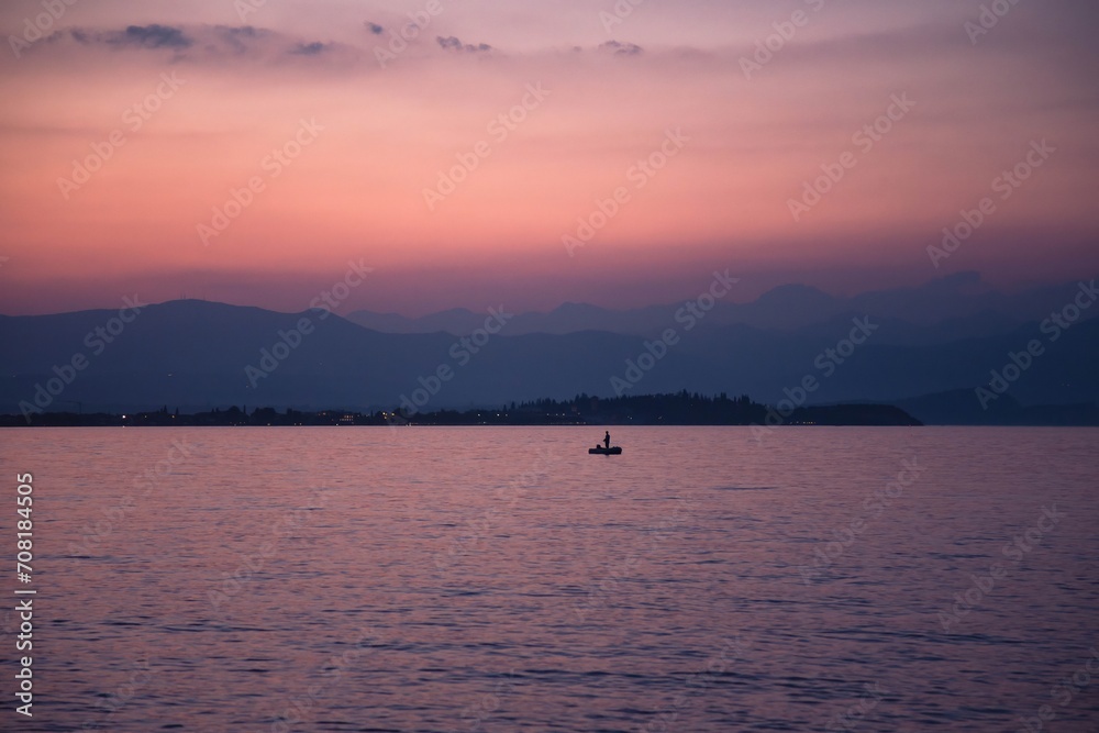 A lonely fisher standing on an inflatable boat with an oar against a backdrop of misty mountains and a Garda lake A lonely fisher standing on an inflatable boat with an oar against a backdrop of misty mountains and a Garda lake