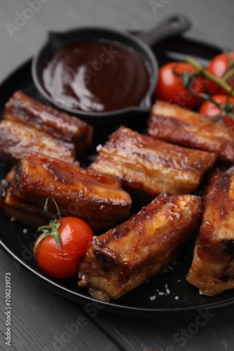 Tasty roasted pork ribs served with sauce and tomatoes on grey wooden table, closeup