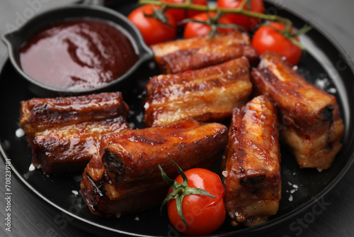 Tasty roasted pork ribs served with sauce and tomatoes on grey wooden table, closeup