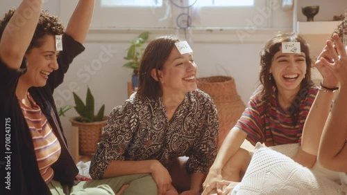 Young joyous girl winning Who Am I game and raising arms in excitement, company of cheerful female friends clapping hands and laughing while hanging out and having fun together at home