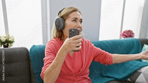 A joyful mature woman enjoys music with headphones and smartphone in her cozy living room. photo