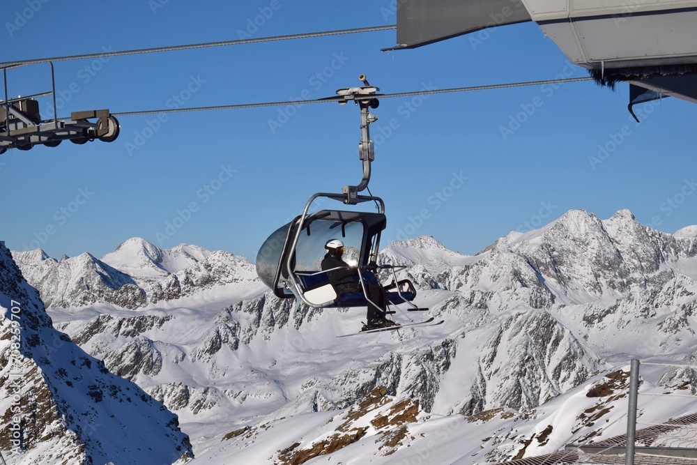 Person sitting in chair lift at the top station of Hochgurgl ski resort, Tyrol, Austria. Overhead lift for skiers and snowboarders to the Top Mountain Star on a sunny day. View of snowy mountain peaks