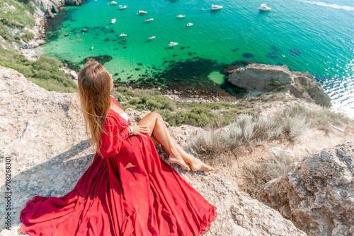 Woman red dress sea. Happy woman in a red dress and white bikini sitting on a rocky outcrop  gazing out at the sea with boats and yachts in the background.