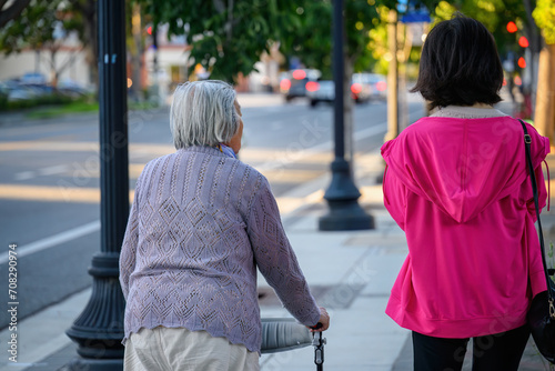 Senior woman walking using a mobility walker on the pedestrian footpath, accompanied by her daughter.