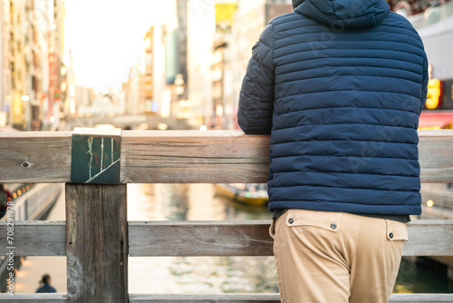 A man wearing blue down jacket is standing and leaning on wooden rail with background of tourist city place. Photo applied with orange sunlight. photo