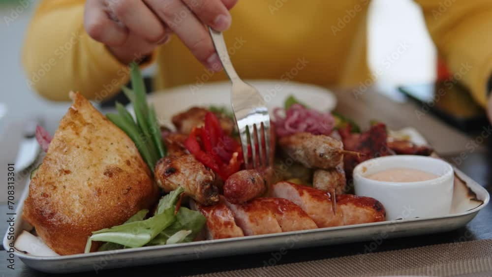 Unrecognizable man spears grilled sausage on fork. Guy eats large platter with various meat and vegetables cooked on barbecue. Heavy fatty foods with lot of calories.