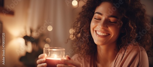 Content woman finds joy in candle scent, smiling at lit candle in cozy home atmosphere at night. Self-care and relaxation.