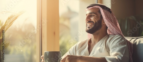 Indian man smiling, drinking hot beverage at home in morning. Arabic guy enjoying alone time, taking a break. photo