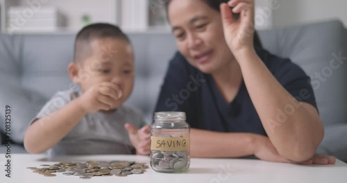 Asian mother with her little 4s son lying at home count and drop coins into glass jar. Caring parent teaches child save money, think about future, manage personal finances, savings concept. photo